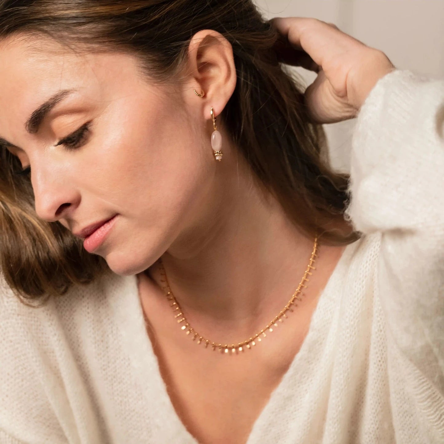 Femme brune portant de fines boucles d'oreilles en or et pierre rose ainsi qu'un collier chaine en or