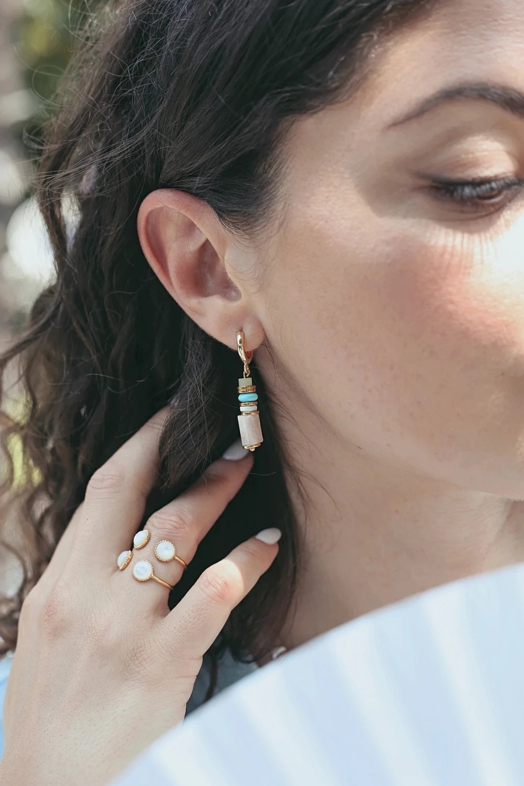 Femme brune portant de fines boucles d'oreilles pendantes colorées pastel et un collier aux grosses pierres pastel