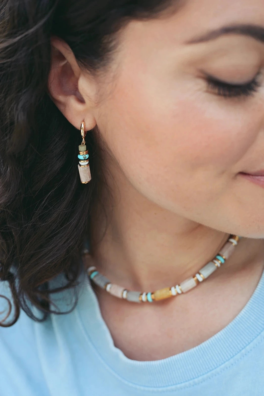 Femme brune portant de fines boucles d'oreilles pendantes colorées pastel et un collier aux grosses pierres pastel